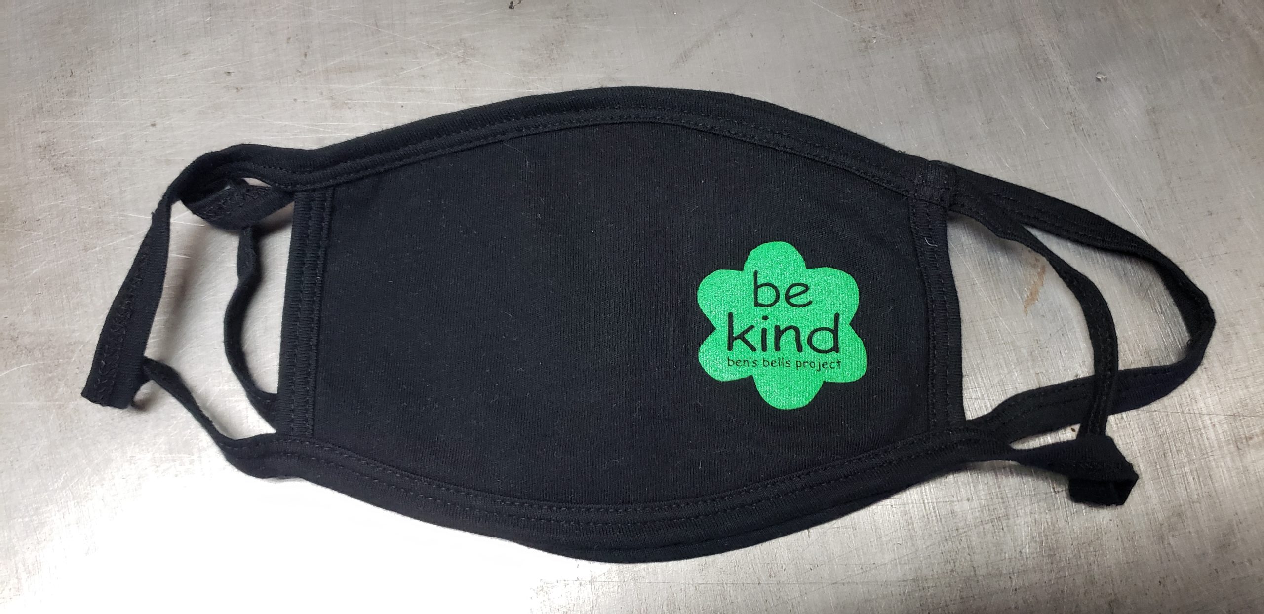 Black cotton face mask with union screen printed one color design for the ben's bells project that reads, "be kind" in a simple flower shape