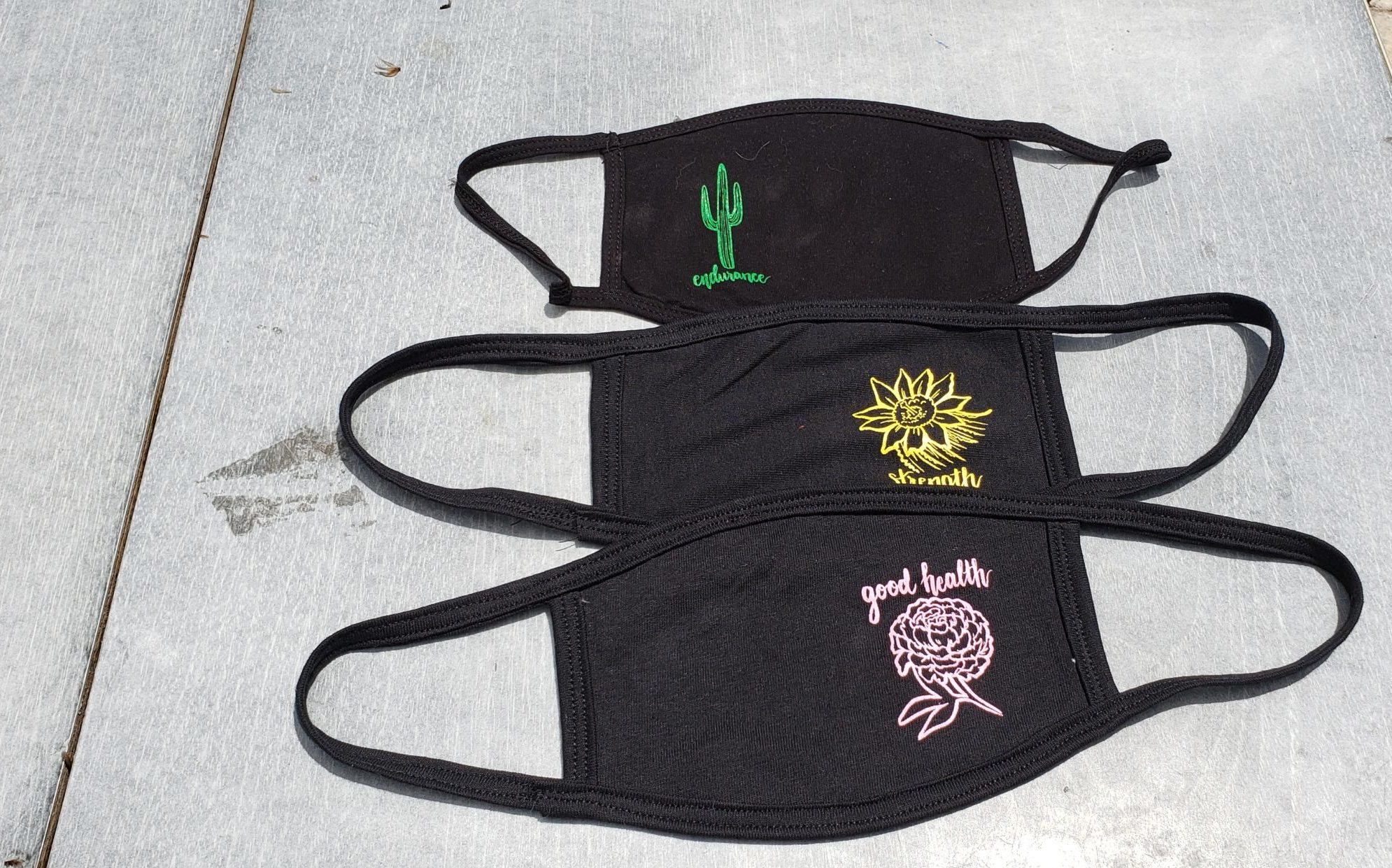 three handmade fair trade black face mask with screen printed designs on the sides of the masks. The three designs from front to back are of a soft pink chrysanthemum with the text, "good health" above it in cursive, a yellow sunflower motif with the word, "Strength" below it, and a green two-limbed saguaro cactus with the word, "endurance" below it.